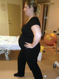 A pregnant woman stands her feet hip width apart and with one foot in front of the other. She has her hands on her hips and her front leg is bent at the knee in a lunging position.