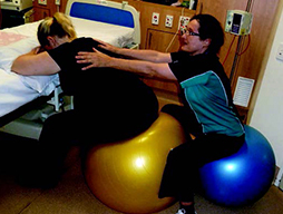 A pregnant woman sits on a fitball facing the side of a a bed. Her folded arms rest on the mattress. Behind the woman sits another person, also on a fitball.