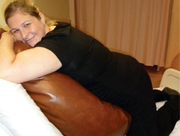 A pregnant woman resting on her knees on a bed leans into a beanbag that is supported by the raised head of the bed.