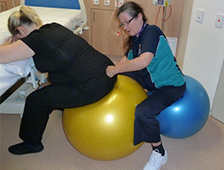 A woman sits on a fitball and leans onto a bed for support. Another women behind her, also seated on a fitball, applies pressure on the base of the first womans's spine with the knuckles of her clenched hand