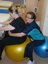 A woman sits on a fitball and leans onto a bed for support. Another women behind her, also seated on a fitball, has her arms either side of the first woman and her hands on her abdomen