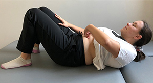 A woman lies on her back with the fingers of one hand at her belly button and rests her other on the thigh of the same leg.