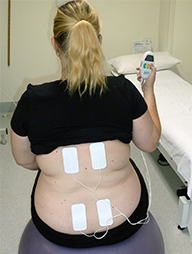A woman faces away from the camera with her t-short lifted up on her back and holding a small device in her right hand.  The device is connected to four adhesive strips placed on her back.