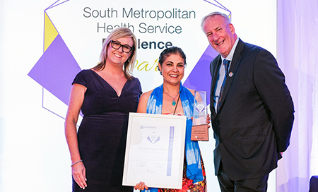 Two women and a man stand in front of a banner that reads South Metropolitan Health Service Excellence Awards. One woman holds a framed certificate.
